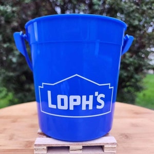 Puede incluir: Un cubo azul brillante con el texto blanco "LOPH'S". El cubo tiene un asa a cada lado y se apoya en una pequeña paleta de madera. El fondo está borroso, con follaje verde.