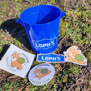 Peut inclure: Un seau bleu avec le texte "LOPH'S" et plusieurs autocollants. Les autocollants présentent un corgi de dessin animé avec une coquille verte et le texte "Chicago Cactus Club". Les articles sont sur une surface herbeuse.