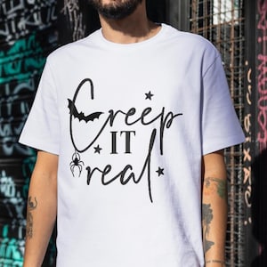 Könnte beinhalten: Weißes T-Shirt mit dem Schriftzug "Creep It Real" in schwarzer Schrift, mit Fledermaus, Spinne und Sternen. Ein lässiges Oberteil, passend für Halloween.