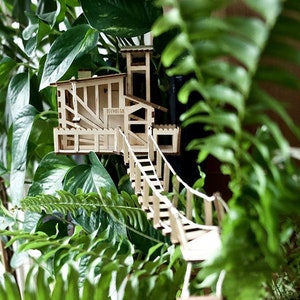 Op de afbeelding: Een houten boomhutmodel met een touwbrug en een ladder, omringd door groene planten in potten.