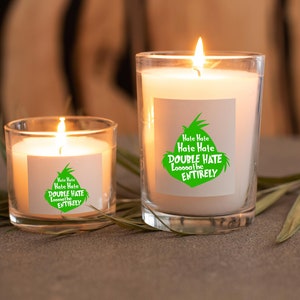 May include: Two lit candles in clear glass jars. The candles have white wax and are labeled with green text that reads "Hate Hate, Hate Hate, DOUBLE HATE Loooooathe ENTIRELY". The candles are on a gray surface.