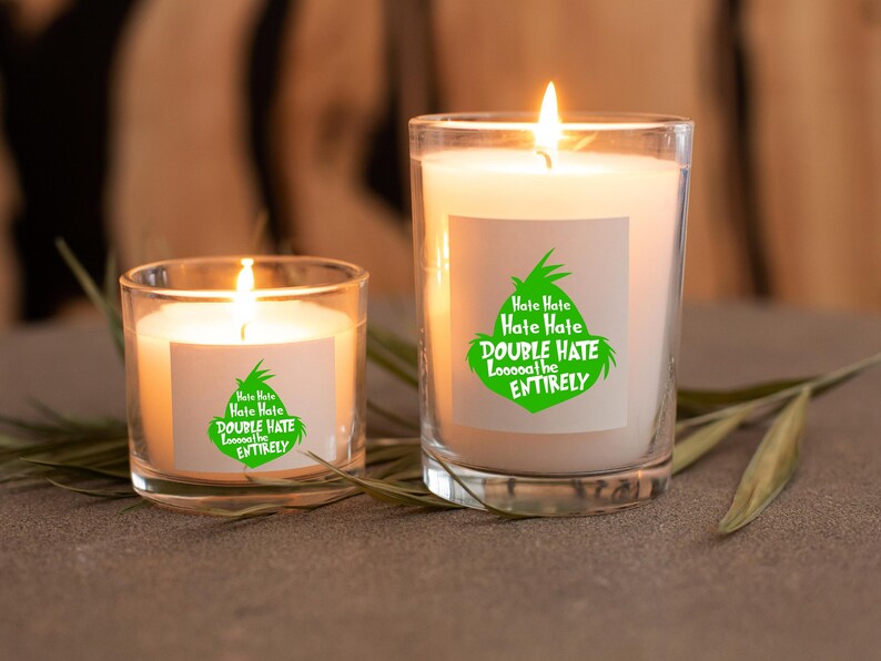 May include: Two lit candles in clear glass jars. The candles have white wax and are labeled with green text that reads "Hate Hate, Hate Hate, DOUBLE HATE Loooooathe ENTIRELY". The candles are on a gray surface.