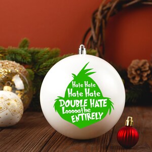 May include: White Christmas ornament with a green Grinch design and the text "Hate Hate Hate Hate Double Hate Looooathe Entirely". The ornament is surrounded by other Christmas decorations.