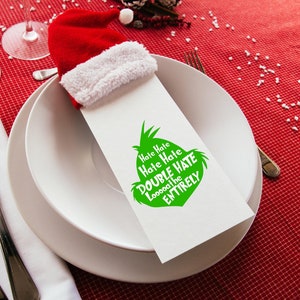 May include: A festive holiday table setting with a red and white Santa hat napkin holder. The white napkin has a green graphic with the text "Hate Hate Hate Double Hate Loooooathe the Entirely". The setting includes white plates, silverware, and wine glasses.