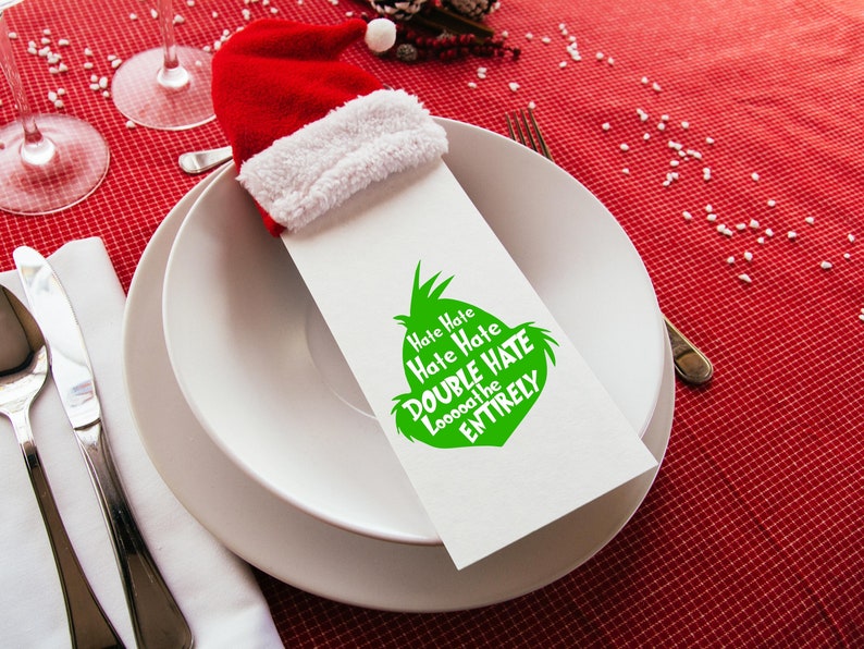 May include: A festive holiday table setting with a red and white Santa hat napkin holder. The white napkin has a green graphic with the text "Hate Hate Hate Double Hate Loooooathe the Entirely". The setting includes white plates, silverware, and wine glasses.