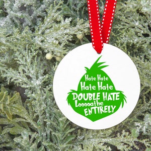 May include: A white, round Christmas ornament with a green Grinch-inspired design and the text "Hate Hate, Double Hate, Looooathe ENTIRELY". The ornament hangs from a red ribbon against a backdrop of frosted greenery.