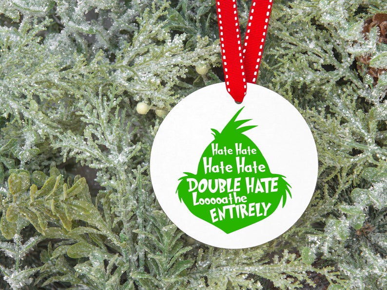 May include: A white, round Christmas ornament with a green Grinch-inspired design and the text "Hate Hate, Double Hate, Looooathe ENTIRELY". The ornament hangs from a red ribbon against a backdrop of frosted greenery.