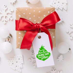 May include: A wrapped gift with a red satin bow and a white tag. The tag features a green Grinch-like character and the text "Hate Hate Hate, Double Hate, Loooooathe ENTIRELY". The gift is on a white surface with Christmas decorations.