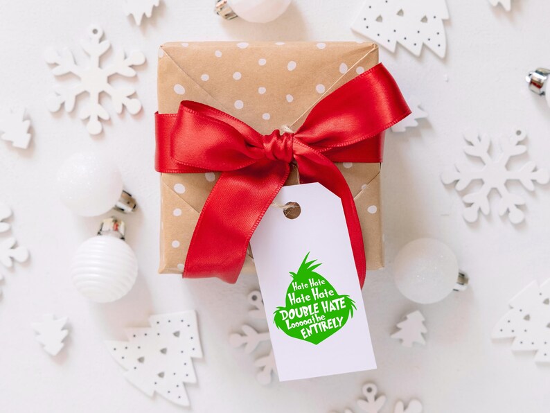 May include: A wrapped gift with a red satin bow and a white tag. The tag features a green Grinch-like character and the text "Hate Hate Hate, Double Hate, Loooooathe ENTIRELY". The gift is on a white surface with Christmas decorations.