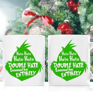 May include: Two white ceramic mugs with a green graphic of a character's face and the text "Hate Hate, Hate Hate, DOUBLE HATE, Looooathe, ENTIRELY". The mugs are in front of a Christmas tree.