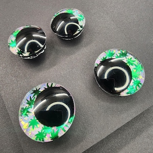 Puede incluir: Cuatro cabujones de resina negros y transparentes con un diseño curvo blanco y acentos de hojas verdes. Los cabujones son ovalados y redondos, con un acabado brillante. El fondo es una superficie gris oscuro.