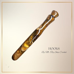 May include: A handcrafted crochet hook with a marbled design in shades of gold, brown, and blue. The hook is angled and features the text "HOOKS By Mr. Toy Shop Crochet".