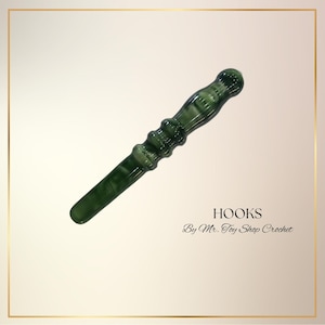 May include: A handcrafted, green crochet hook with a marbled appearance. The hook has a rounded handle and a tapered end. The text "HOOKS By Mr. Toy Shop Crochet" is displayed below the hook.