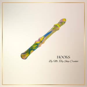 May include: A colorful crochet hook with a marbled design in shades of yellow, blue, and pink. The hook has a textured, ergonomic handle. Text on the image reads "HOOKS By Mr. Toy Shop Crochet."
