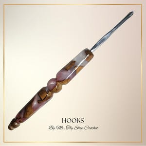 May include: A crochet hook with a silver metal hook and a handle made of pink, brown, and clear resin. The handle has a bulbous shape with a textured appearance. The text "HOOKS By Mr. Toy Shop Crochet" is at the bottom.