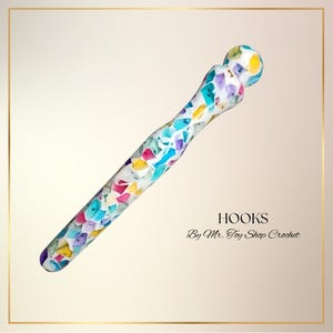 May include: A colorful crochet hook with a mosaic pattern in shades of blue, purple, yellow, and pink. The hook has a rounded end and a textured shaft. Text on the image reads "HOOKS By Mr. Toy Shop Crochet."