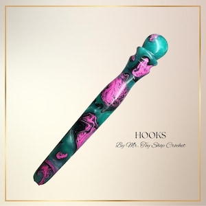 May include: A crochet hook with a teal, pink, and black marbled design. The hook has a rounded top and a textured handle. The text "HOOKS By Mr. Toy Shop Crochet" is visible.
