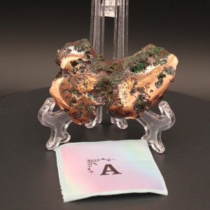 May include: A large, irregular piece of copper ore with a rough, textured surface. The ore is a mix of copper and green malachite, and is displayed on a clear plastic stand. The ore is labeled with the letter "A".