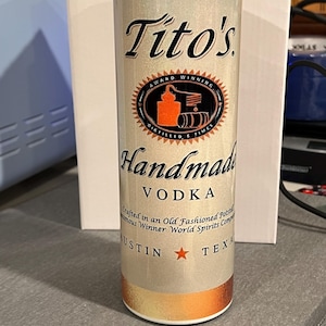 Pu&ograve; includere: Un bicchiere metallico argentato e dorato con sfondo bianco e testo nero. Il testo recita "Tito's Award Winning Distilled 6 Times Handmade Vodka Crafted in an Old Fashioned Pot Still Numerous Winner World Spirits Competition Austin Texas".