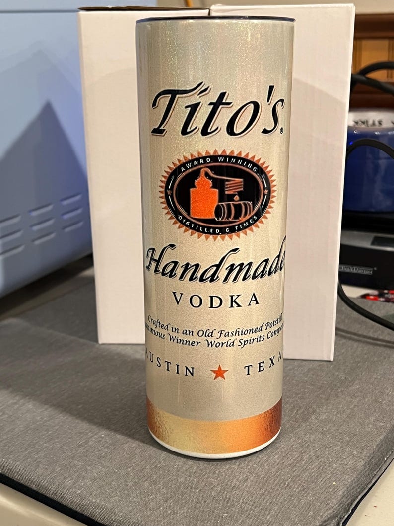 Pu&ograve; includere: Un bicchiere metallico argentato e dorato con sfondo bianco e testo nero. Il testo recita "Tito's Award Winning Distilled 6 Times Handmade Vodka Crafted in an Old Fashioned Pot Still Numerous Winner World Spirits Competition Austin Texas".