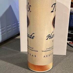 Pu&ograve; includere: Un bicchiere in acciaio inossidabile bianco e oro con una finitura scintillante. Il bicchiere presenta un logo in bianco e nero con il testo "T.I. Handmade Vodka" e "Crafted in Austin, Texas".
