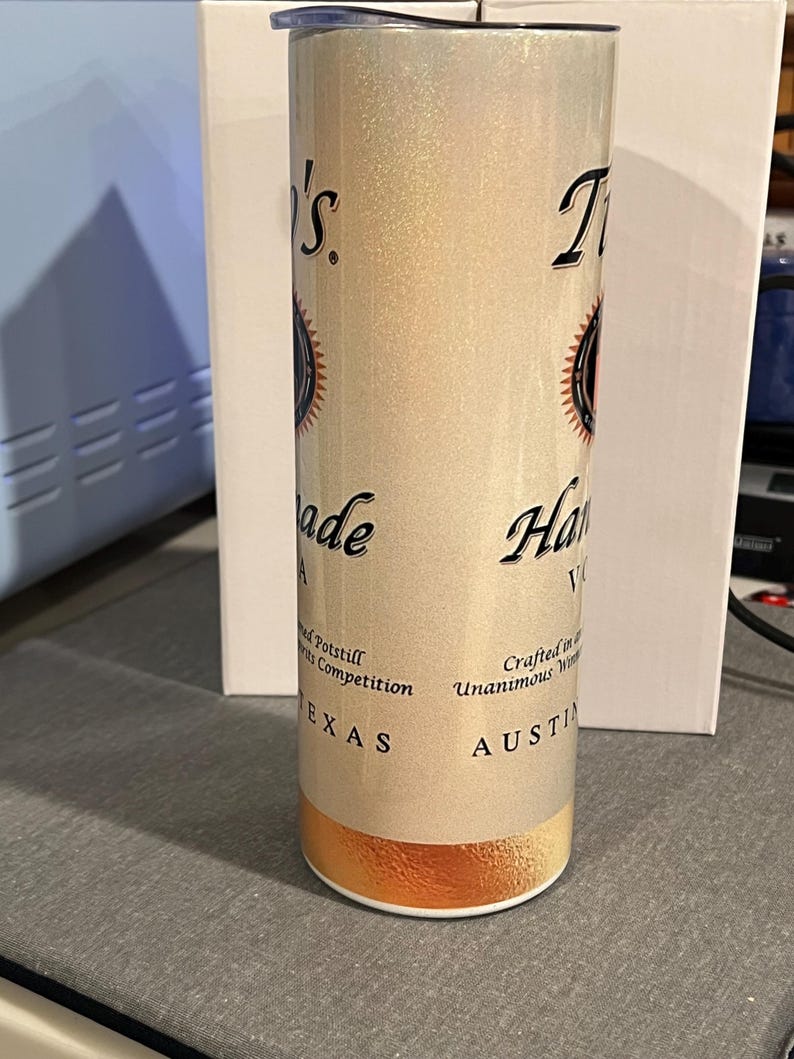 Pu&ograve; includere: Un bicchiere in acciaio inossidabile bianco e oro con una finitura scintillante. Il bicchiere presenta un logo in bianco e nero con il testo "T.I. Handmade Vodka" e "Crafted in Austin, Texas".
