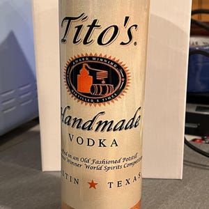 Pu&ograve; includere: Un bicchiere in acciaio inossidabile bianco e oro con un logo Tito's Handmade Vodka. Il logo presenta un'illustrazione in nero e arancione di un alambicco e il testo "Award Winning" e "Distilled 6 Times". Il bicchiere presenta anche il testo "Handmade Vodka" e "Austin, Texas".