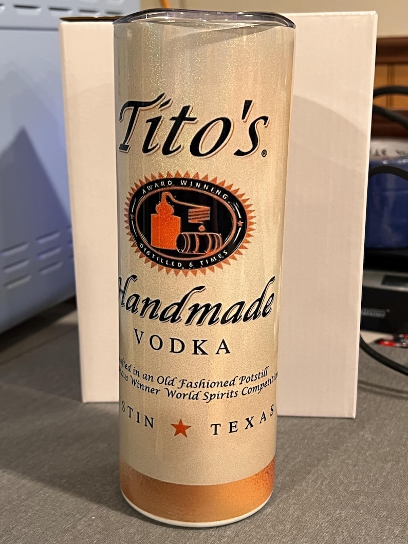 Pu&ograve; includere: Un bicchiere in acciaio inossidabile bianco e oro con un logo Tito's Handmade Vodka. Il logo presenta un'illustrazione in nero e arancione di un alambicco e il testo "Award Winning" e "Distilled 6 Times". Il bicchiere presenta anche il testo "Handmade Vodka" e "Austin, Texas".