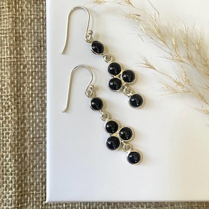 May include: A pair of silver drop earrings featuring a vertical arrangement of small, round, polished black stones. Each earring consists of a hook wire connected to a single stone, followed by a cluster of three stones and a final bottom stone.