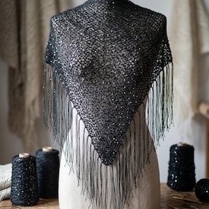 Puede incluir: Un chal negro triangular con un tejido delicado y abierto y lentejuelas brillantes. Largas borlas cuelgan del borde inferior. El chal se exhibe en un maniquí. Bobinas de hilo negro en primer plano.