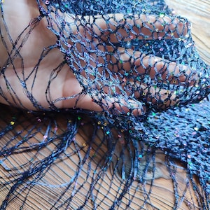 May include: Close-up of a navy blue, open-weave fabric with iridescent sequins. The fabric has a net-like structure with long, fringed edges. The sequins catch the light, creating a shimmering effect. The fabric is likely used for apparel or decorative purposes.