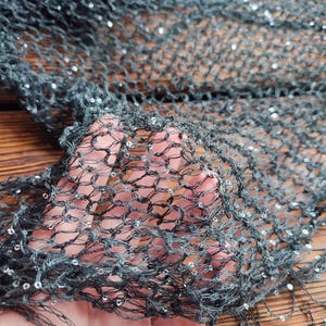 May include: Close-up of a dark gray, open-weave fabric with small, reflective sequins. The fabric has a net-like appearance, with visible knots and a slightly frayed edge. The material is held up by a hand.