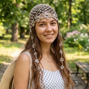 May include: A beige crochet skull cap featuring a diamond-shaped mesh pattern, embellished with small metallic sequins. The cap includes floral-shaped accents along the front edge and two long, dangling side strands adorned with matching floral motifs.