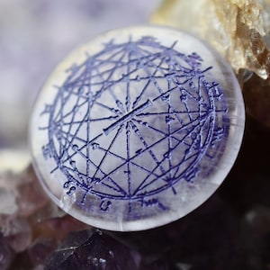 Könnte beinhalten: Eine klare Glaskugel mit einem eingeätzten violetten geometrischen Muster auf der Oberfläche. Die Kugel liegt auf einem Bett aus Amethystkristallen.