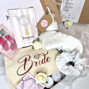Könnte beinhalten: Eine Geschenkbox gefüllt mit Artikeln für eine zukünftige Braut. Die Box enthält eine weiße Satin-Schlafmaske, ein weißes Satin-Haargummi, eine weiße Augenmaske, eine kleine Tasche mit dem Wort "Bride" darauf, eine Tüte Gummibärchen, eine Tüte Tee und eine kleine weiße Box mit Kerzen.