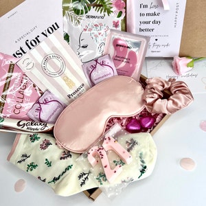 May include: A gift box containing a pink satin sleep mask, a matching scrunchie, cream-colored socks with a floral pattern, a number 21 candle, a Galaxy Ripple chocolate bar, Prosecco gummies, Pukka tea bags, and various skincare face masks.