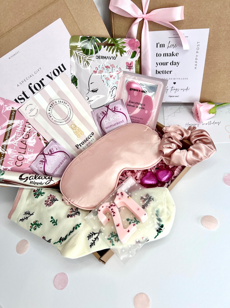 May include: A gift box containing a pink satin sleep mask, a matching scrunchie, cream-colored socks with a floral pattern, a number 21 candle, a Galaxy Ripple chocolate bar, Prosecco gummies, Pukka tea bags, and various skincare face masks.