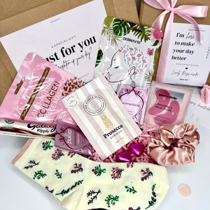 May include: A pink and white gift box filled with various items, including a face mask, a pink hair scrunchie, a bag of gummies, and a pair of socks with a floral pattern. The box is decorated with a pink ribbon and a card that says "I'm here to make your day better".