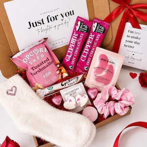 May include: A gift box containing a pair of white fuzzy socks with a heart, two pink Clipper hot chocolate sachets, a bag of Popcorn Shed toasted marshmallow popcorn, a Galaxy Ripple bar, pink eye masks, heart-shaped candies, and a pink scrunchie.