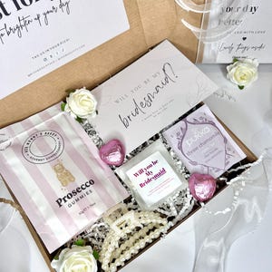 May include: A bridesmaid gift box containing a card, Prosecco gummies, a hair clip, and tea. The box is decorated with white flowers and ribbon. The Prosecco gummies weigh 50g.