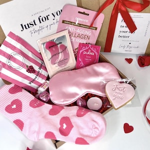 May include: A pink and white gift box filled with self-care items, including a pink eye mask, a pink face mask, a pink sleep mask, a pink cookie, a pink and white striped bag of gummies, and a pair of pink socks with red hearts. The box is tied with a red ribbon and has a white tag that says "Just for you".