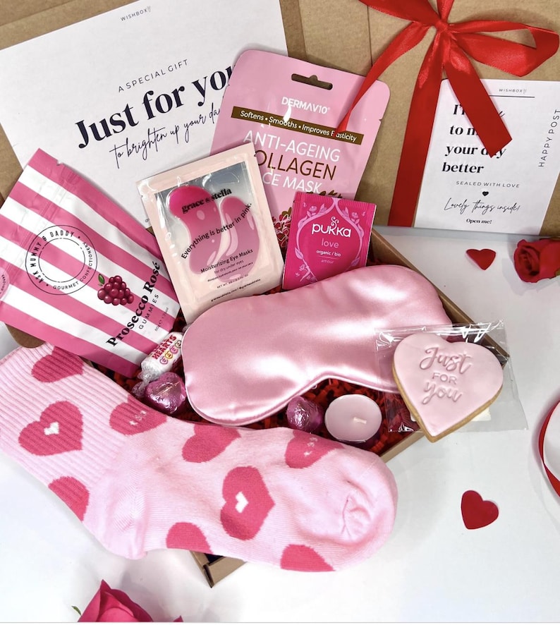 May include: A pink and white gift box filled with self-care items, including a pink eye mask, a pink face mask, a pink sleep mask, a pink cookie, a pink and white striped bag of gummies, and a pair of pink socks with red hearts. The box is tied with a red ribbon and has a white tag that says "Just for you".