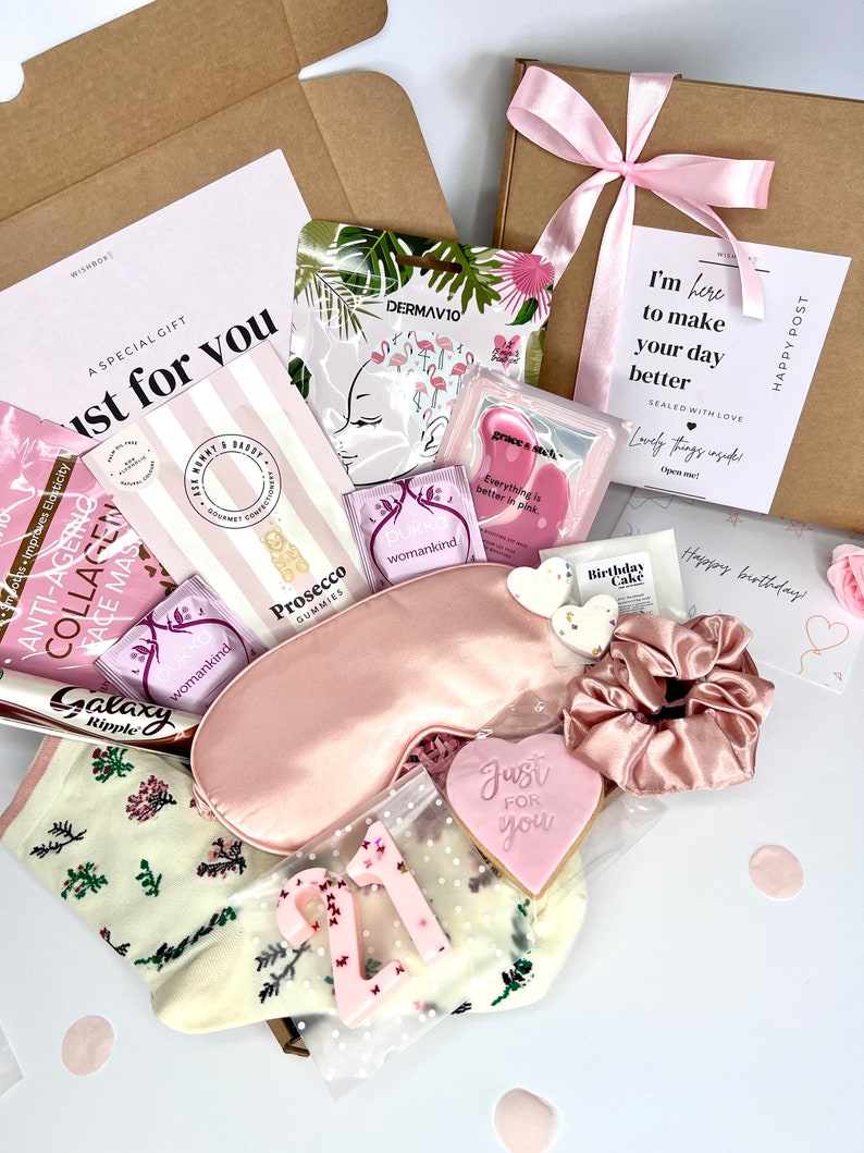 May include: A gift box containing a pink satin sleep mask, a pink scrunchie, a heart-shaped cookie with 'Just for you' text, a '21' candle, Prosecco gummies, collagen face mask, tea bags, a Galaxy Ripple chocolate bar, and floral socks.
