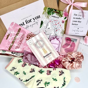 May include: A gift box containing a variety of items including a pair of cream-colored socks with a floral pattern, a pink satin scrunchie, a pack of Prosecco gummies, two Pukka tea bags, a Galaxy Ripple chocolate bar, and several skincare masks.