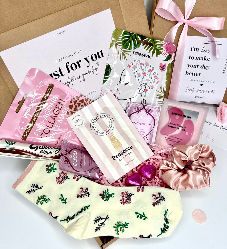 May include: A gift box containing a variety of items including a pair of cream-colored socks with a floral pattern, a pink satin scrunchie, a pack of Prosecco gummies, two Pukka tea bags, a Galaxy Ripple chocolate bar, and several skincare masks.
