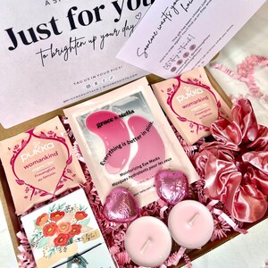 May include: A pink and white gift box filled with self-care items. The box includes eye masks, tea bags, candles, and a small card with a floral design. The box is decorated with pink tissue paper and a pink satin ribbon.