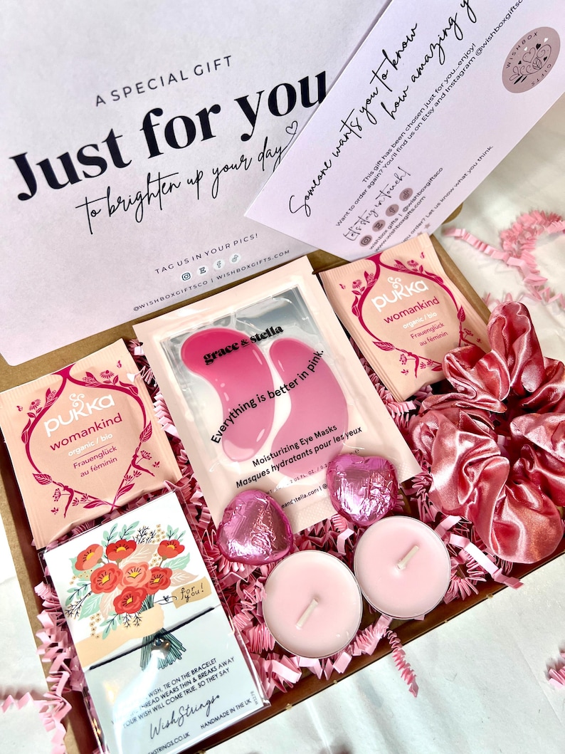 May include: A pink and white gift box filled with self-care items. The box includes eye masks, tea bags, candles, and a small card with a floral design. The box is decorated with pink tissue paper and a pink satin ribbon.