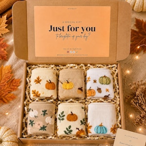 Könnte beinhalten: Eine Geschenkschachtel aus Karton mit sechs Paar gefalteten Socken mit herbstlichen Mustern wie Kürbissen, Blättern und Eicheln. Der Deckel der Schachtel ist mit dem Text "A SPECIAL GIFT Just for you To brighten up your day" und Social-Media-Symbolen bedruckt.