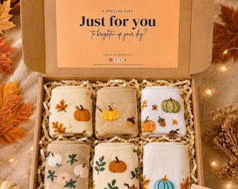 Peut inclure: Une boîte cadeau en carton contenant six paires de chaussettes pliées avec des motifs sur le thème de l'automne, incluant des citrouilles, des glands et des feuilles. Le couvercle porte une carte avec le texte Just for you to brighten up your day.