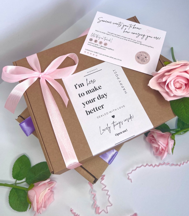 May include: A brown cardboard box with a pink ribbon and a white card that says "I'm here to make your day better. Sealed with love. Lovely things inside. Open me!" The box is surrounded by pink roses.